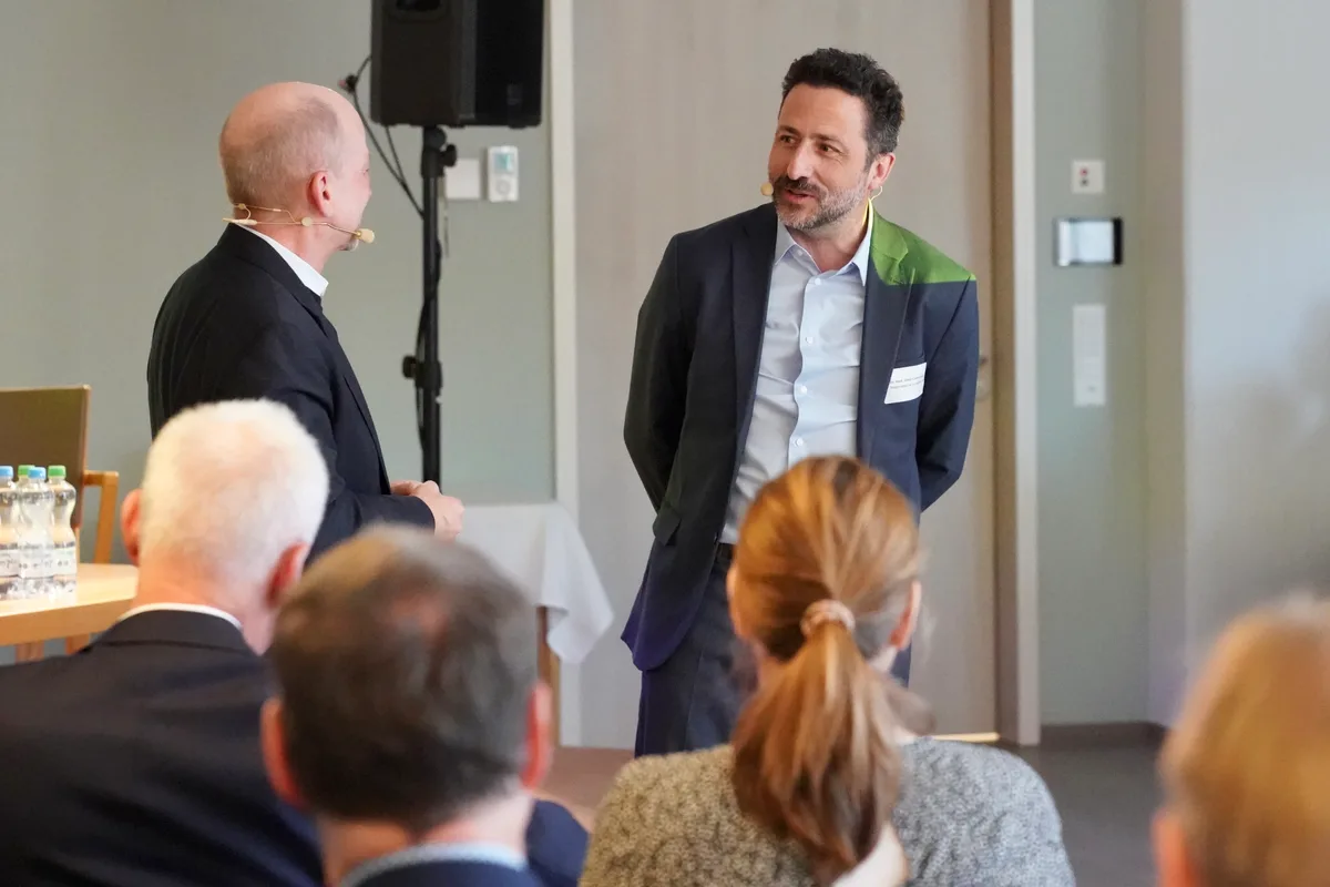 Auf einem Podium stehen zwei Männer, die mit einem Mikrofon verkabelt sind. 